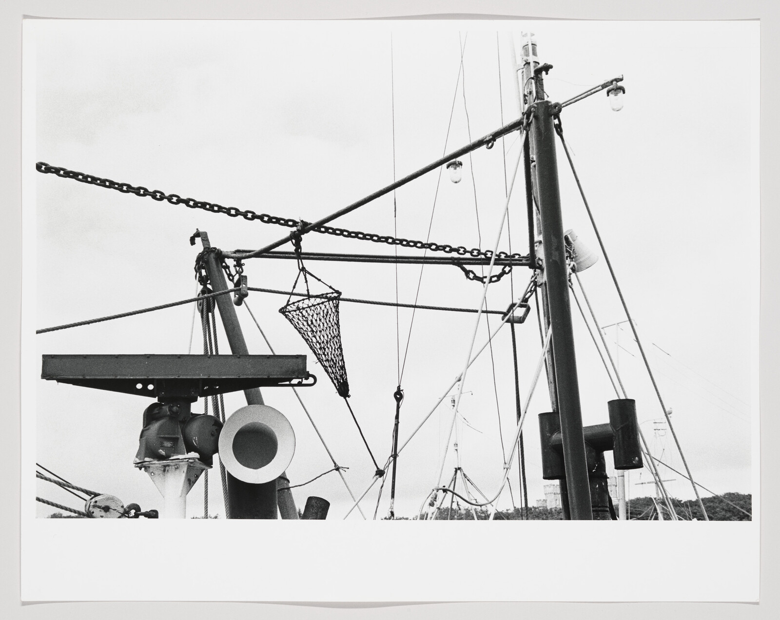Fishing boat rigging with a hanging conical net, chains, and support poles against sky.