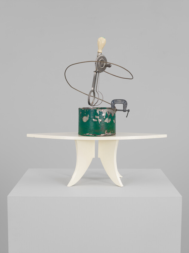 A vintage paint can with an eggbeater and wire loop clamped atop a small white pedestal table.