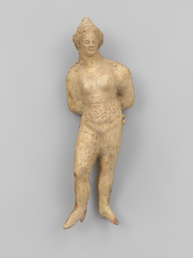 A standing ancient Greek terracotta figurine of a woman, depicted with stylized hair and facial features, wearing a headdress and detailed garments, against a plain gray background.