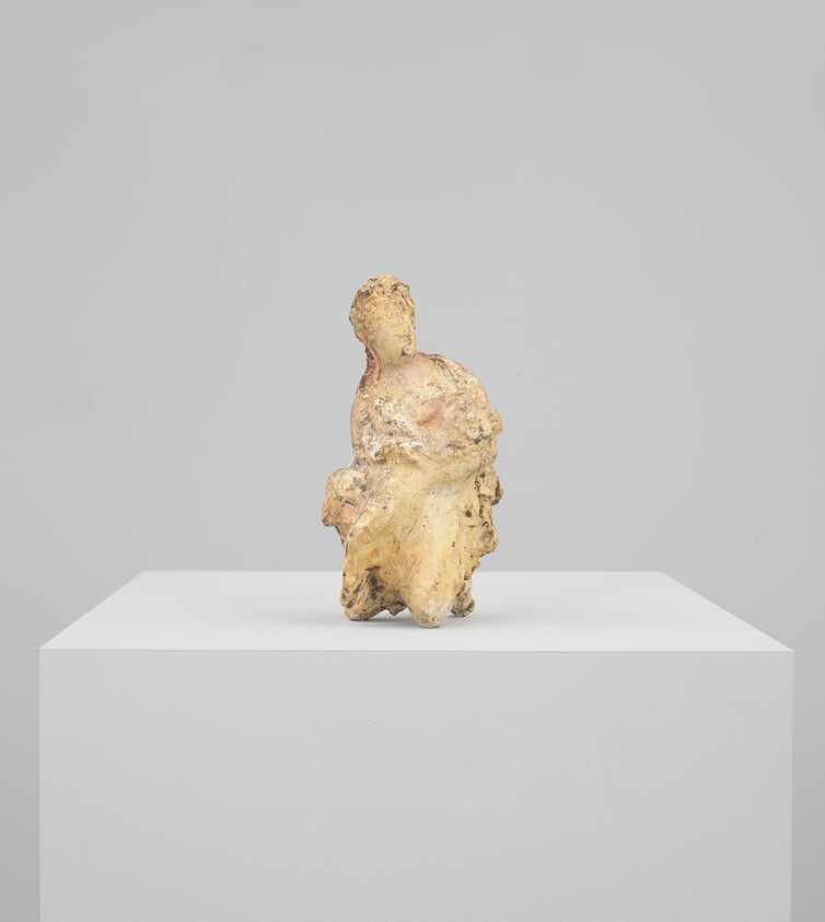 Small worn clay figurine of a seated figure displayed on a white pedestal.