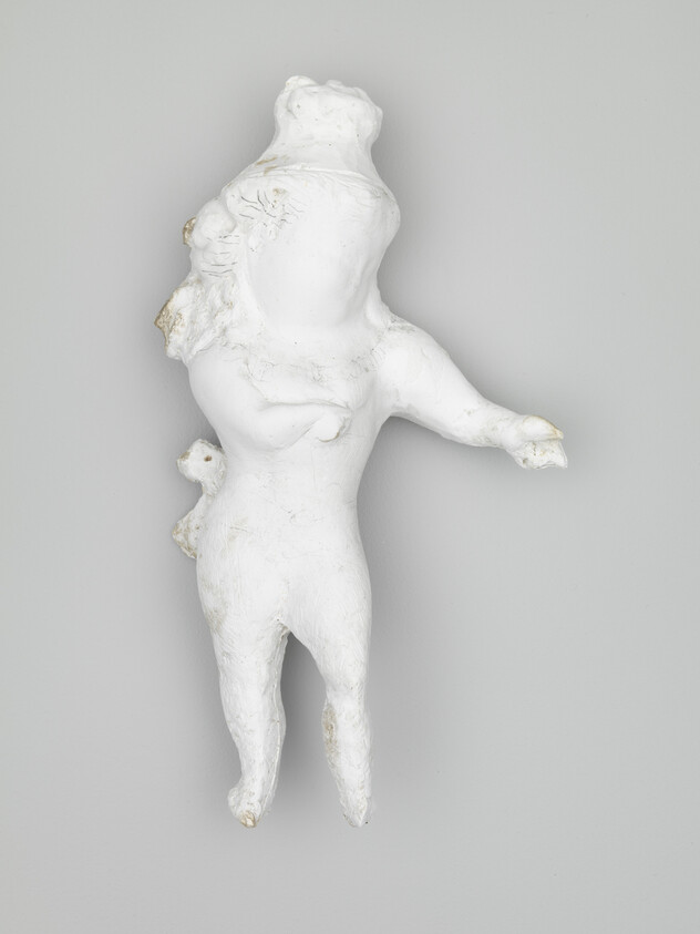 A small white plaster humanoid sculpture with one arm extended and rounded head mounted on a gray wall.