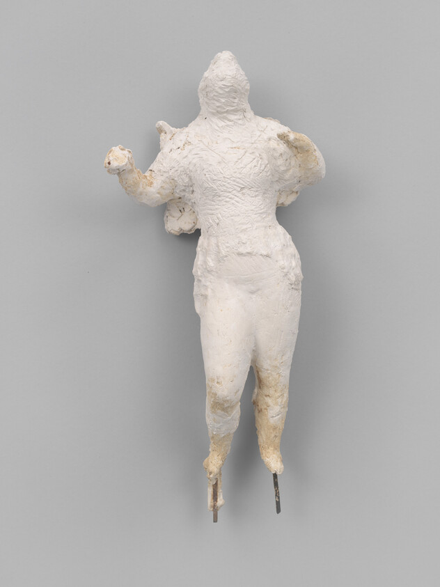 A rough white plaster sculpture of a human figure with arms raised and metal rods protruding from its feet.