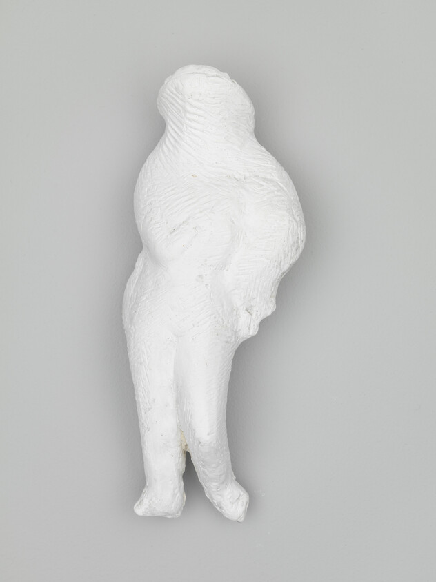 A small white abstract humanoid sculpture with rounded form and merged legs on a gray wall.
