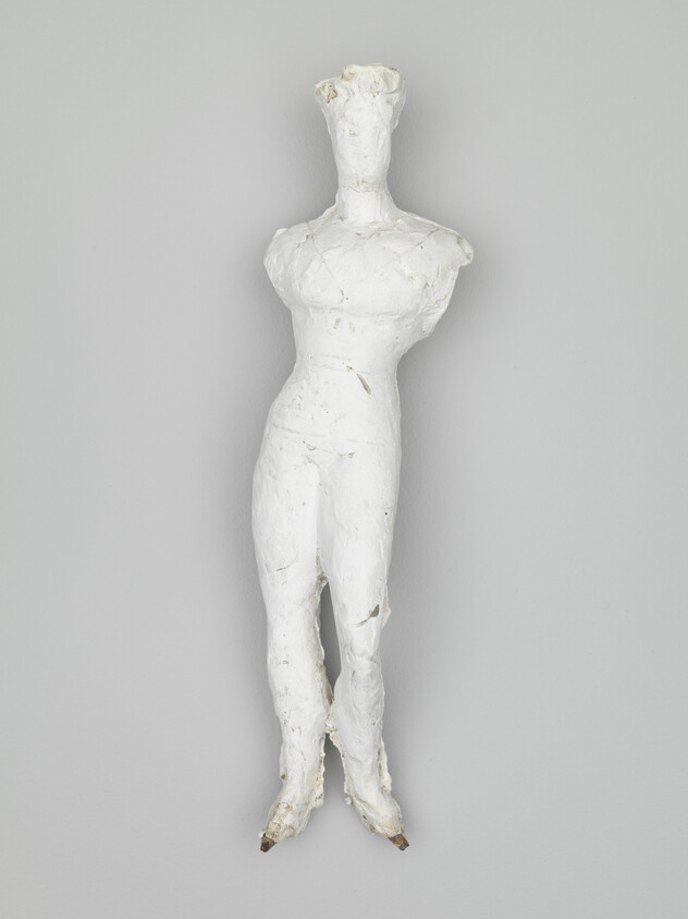 A textured white sculpture of a human figure without arms and a featureless face is mounted on a grey wall.
