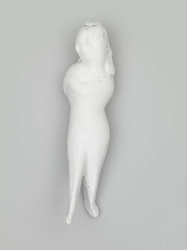 A small white plaster figurine of a standing woman mounted on a plain gray wall.