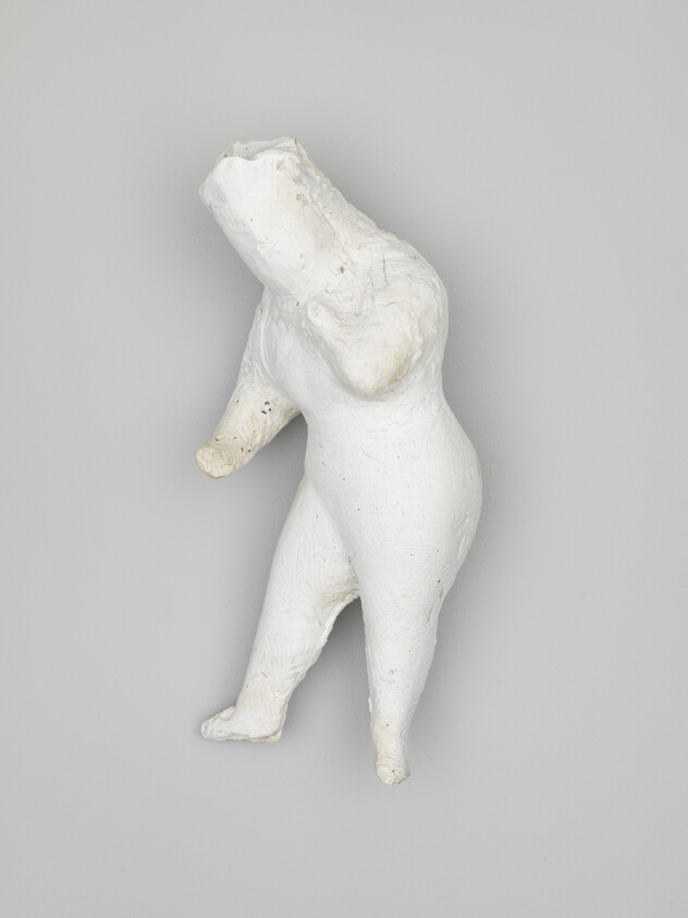 A white plaster abstract torso with missing head and one arm mounted on a pale gray wall.
