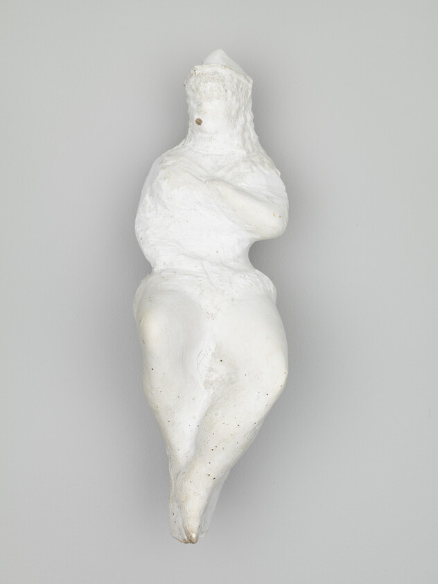 White abstract clay figure with crossed arms and rounded hips mounted on a gray wall.