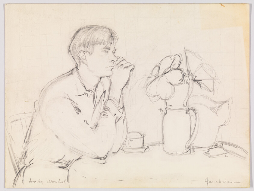 Young man seated at a table, hands clasped near his mouth, looking toward a pitcher of flowers.