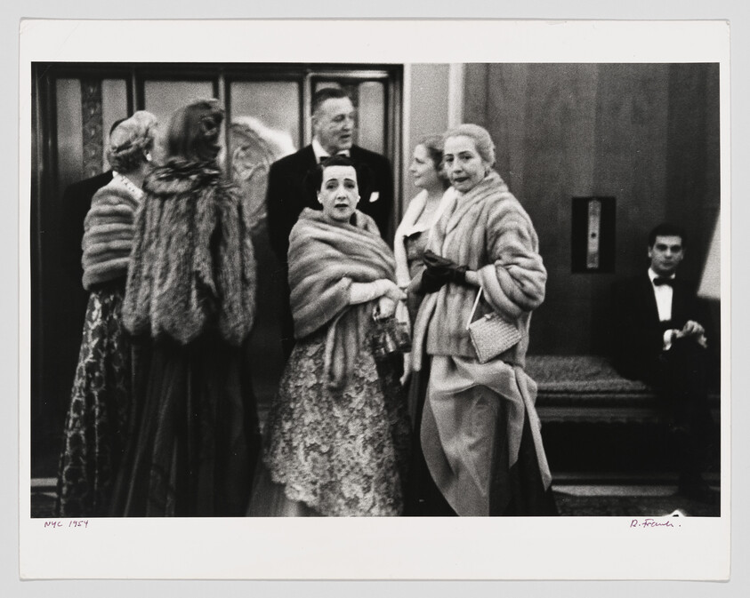 Well-dressed people in evening gowns and fur stoles gather in a lobby, some looking toward camera.