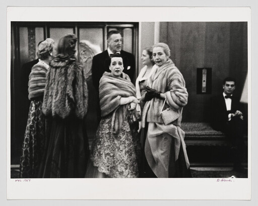 Well-dressed people in evening gowns and fur stoles gather in a lobby, some looking toward camera.