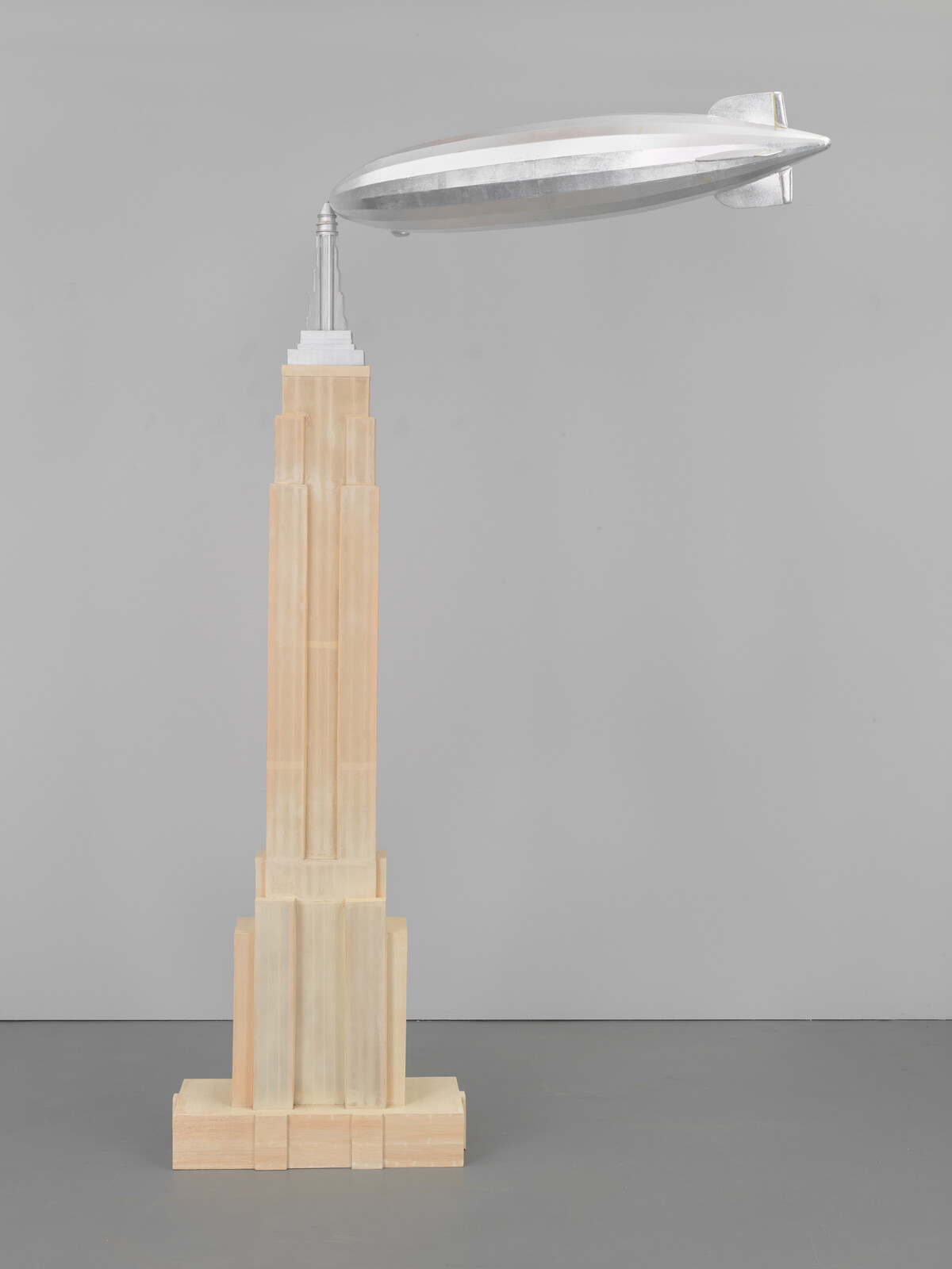 A tall beige model skyscraper topped by a silver blimp floating near its spire.