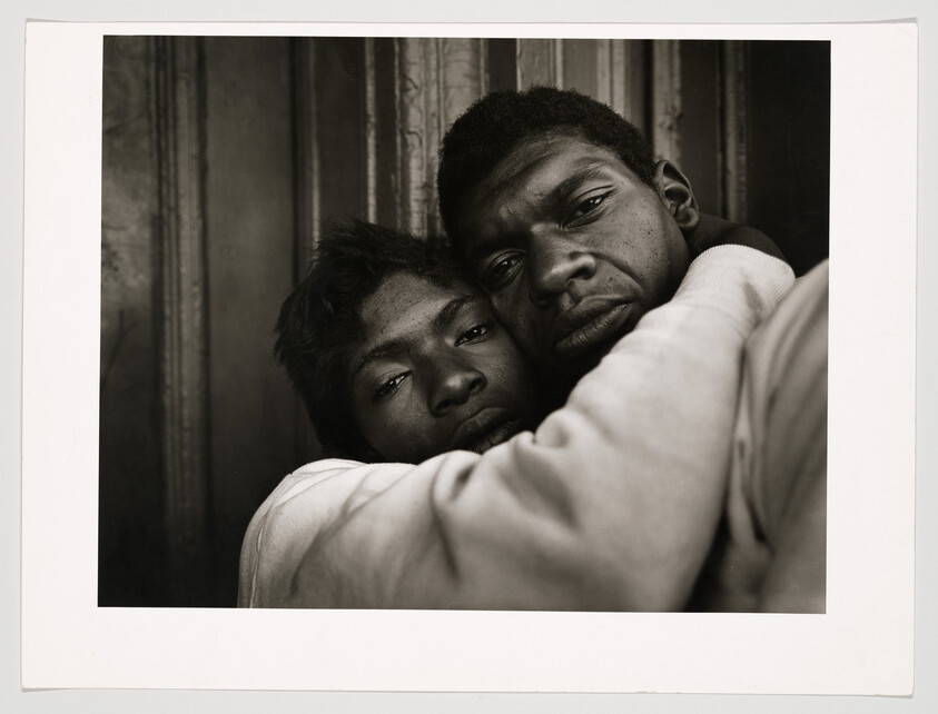 Two men hold each other closely against a corrugated wall, their faces showing quiet weariness.