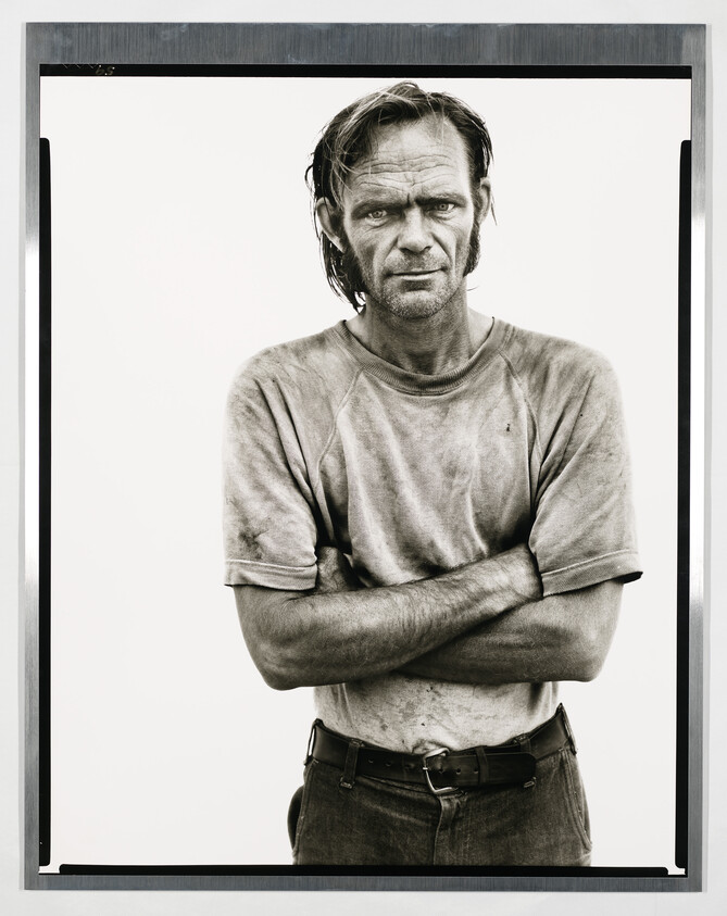 A gritty man stands with arms crossed, wearing a dirty shirt and belt.