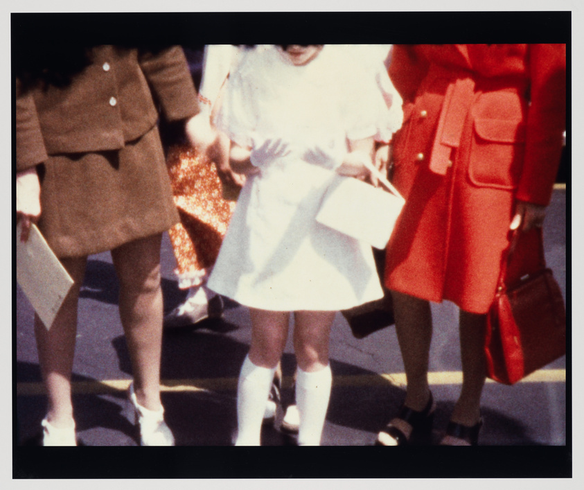 A vintage, overexposed photograph capturing the lower half of three individuals, presumably women, walking side by side. The person on the left is wearing a brown skirt suit and white shoes, holding papers. The middle person is in a white dress with a flared skirt and white knee-high socks with white shoes, also holding papers. The person on the right is dressed in a red coat with a matching skirt, black shoes, and carrying a red handbag. The focus is on their attire and the way they are walking, with the background indistinct.