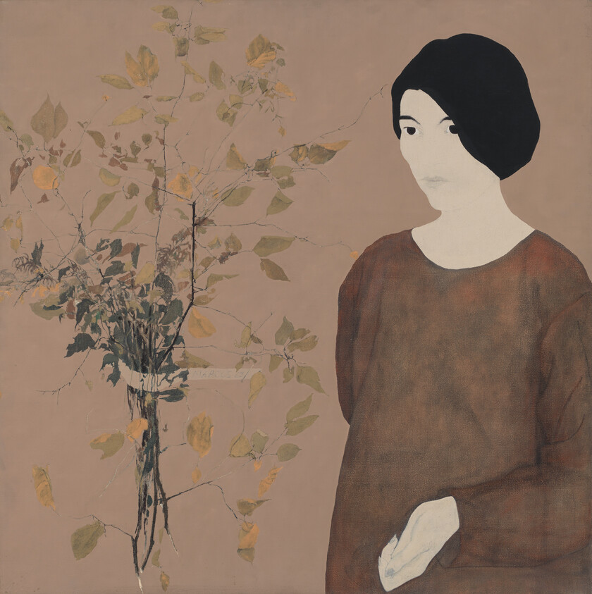 Person with short dark hair stands beside a vase of leafy branches on a muted background.