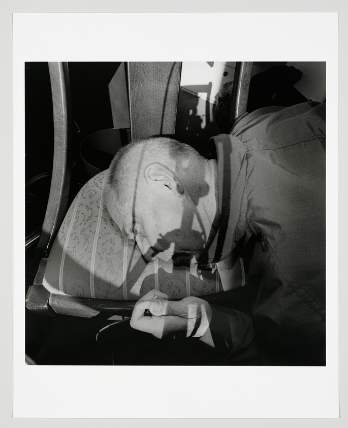 A man rests his head on a patterned chair cushion with his hand clenched.