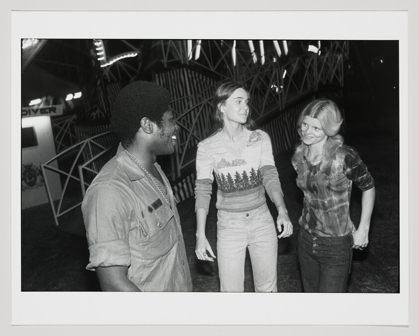 Three young adults chat and laugh together near a lit amusement ride at night.