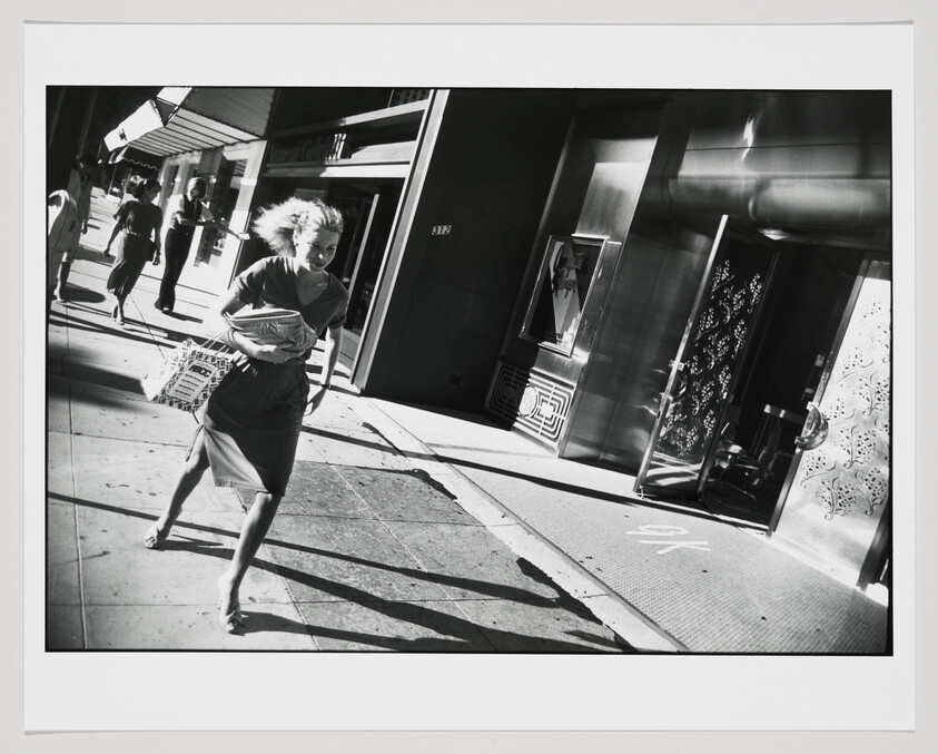 A woman hurries down a city sidewalk holding a bundle of papers as wind blows her hair.
