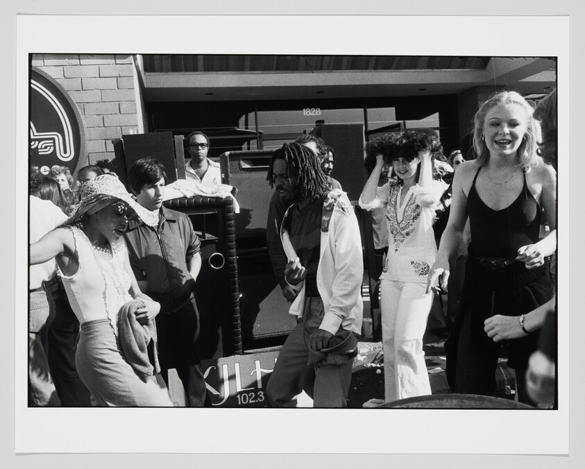 A diverse group of young people dancing and smiling together in front of large speakers outdoors.