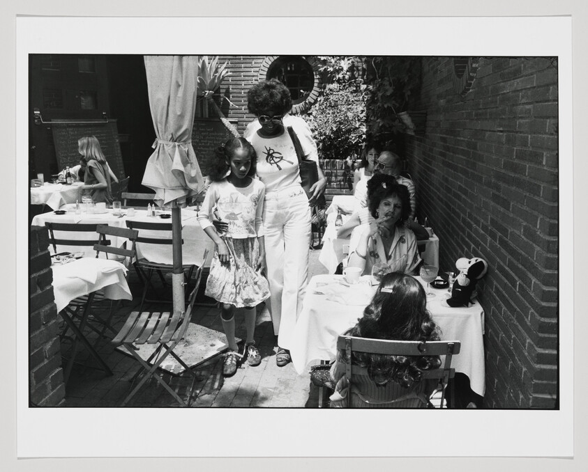 Woman walks arm-in-arm with young girl past café tables while diners sit nearby.
