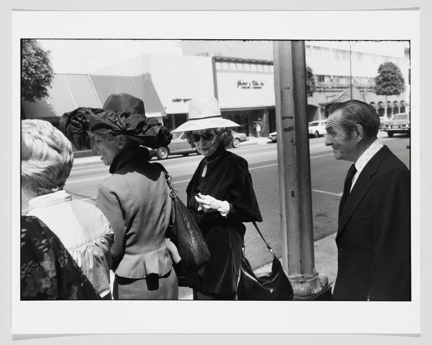 Three women in hats and a man stand on a city sidewalk near a streetlamp.