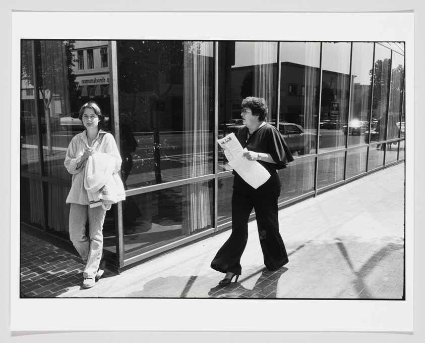 Woman walks briskly holding a newspaper as another woman leans against a building window.