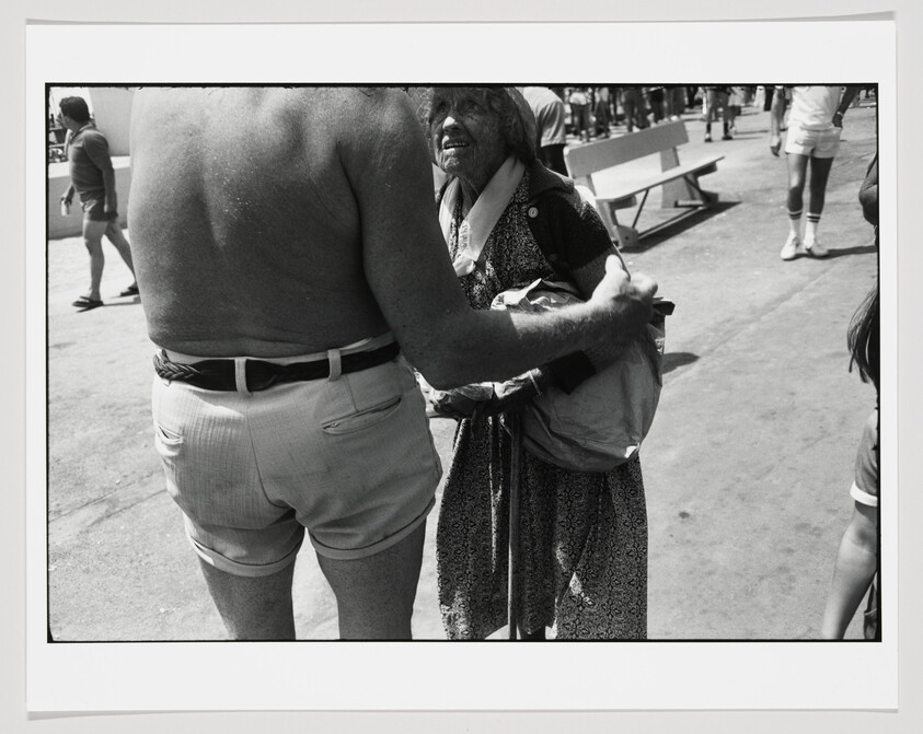 Elderly woman in a patterned dress talks with a shirtless man who gently holds her arm.