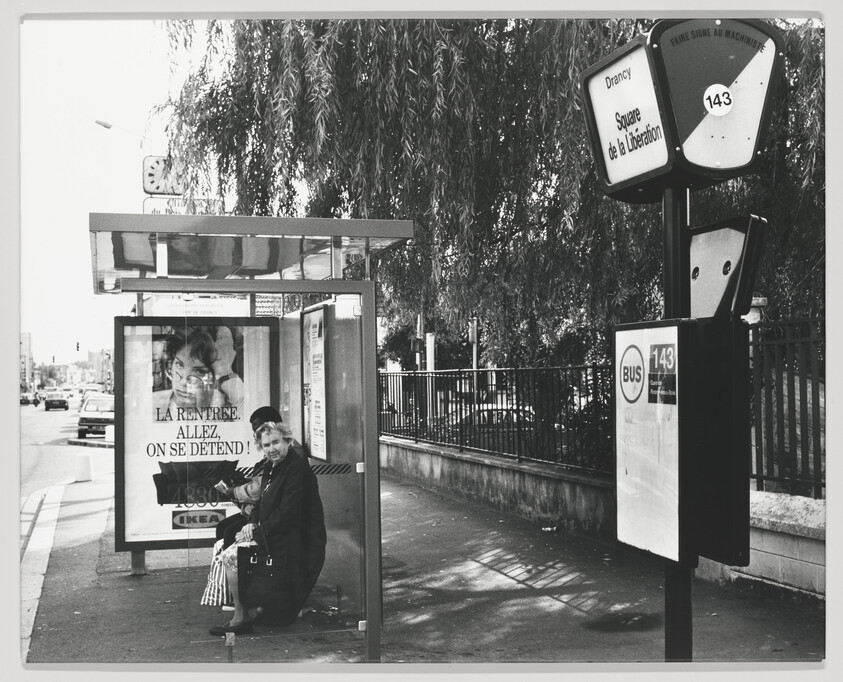 An elderly woman sits at a bus stop shelter near a sign for bus 143.