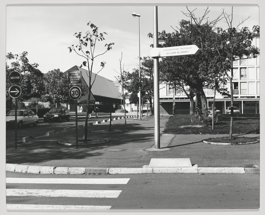 Pedestrian crosswalk leads to a quiet urban intersection with trees, traffic signs, and parked cars.