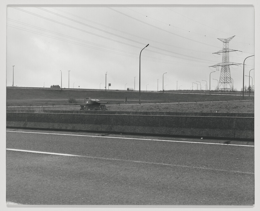 A lone military tank travels on a side road beside a multi-lane highway under power lines.