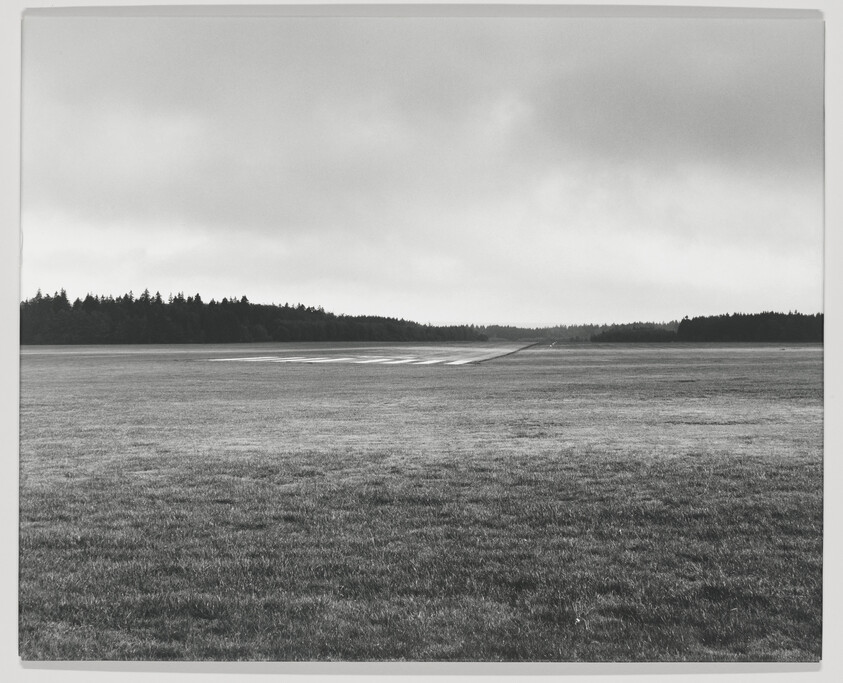 Wide grassy field with a narrow wet runway leading toward a distant tree-lined horizon under cloudy sky.