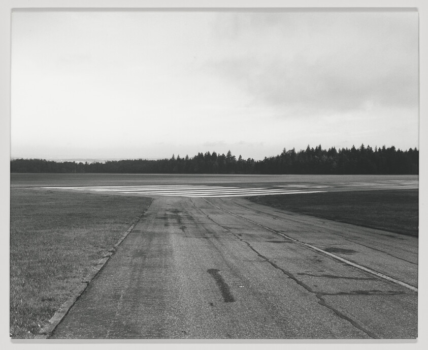 A cracked airport taxiway leads to a striped runway with trees on the horizon.