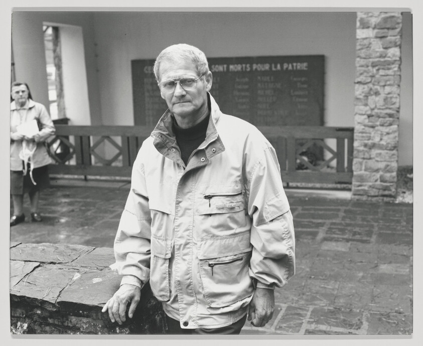 An older man in a light jacket stands solemnly by a war memorial plaque with names.