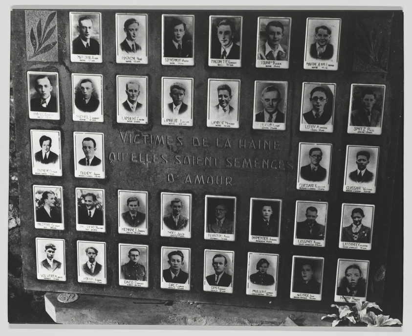 A black and white photo of a memorial wall with multiple framed portraits of individuals. Below the portraits is an inscription in French that translates to "Victims of hatred, may they sow seeds of love." There are flowers placed at the bottom, indicating a site of remembrance.