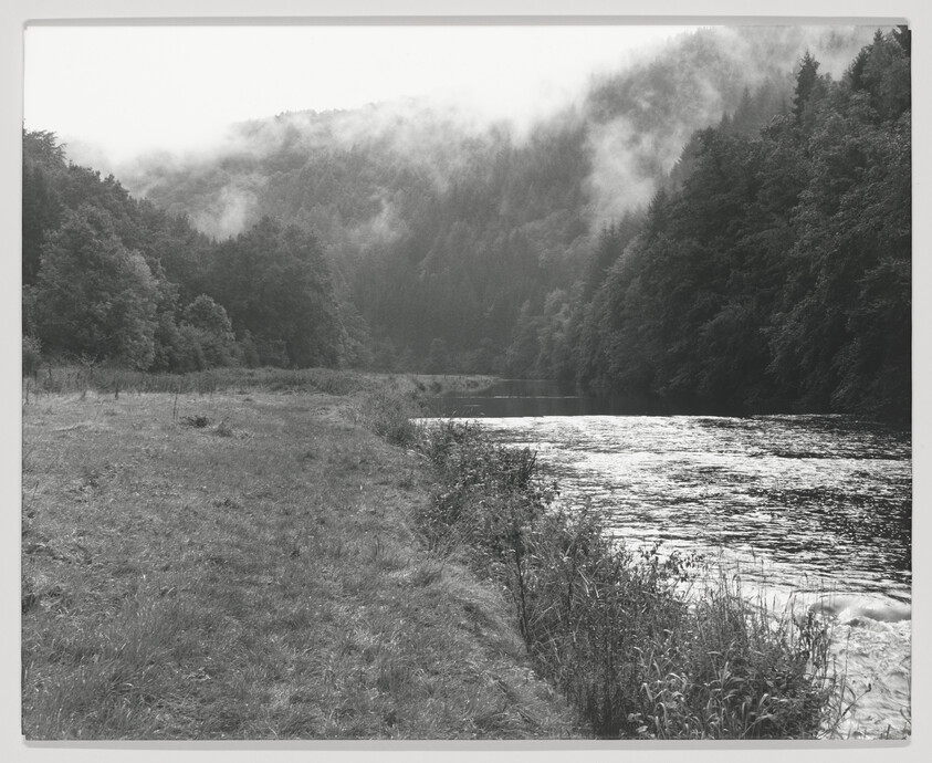 A river runs beside a grassy bank with mist rising over forested hills.