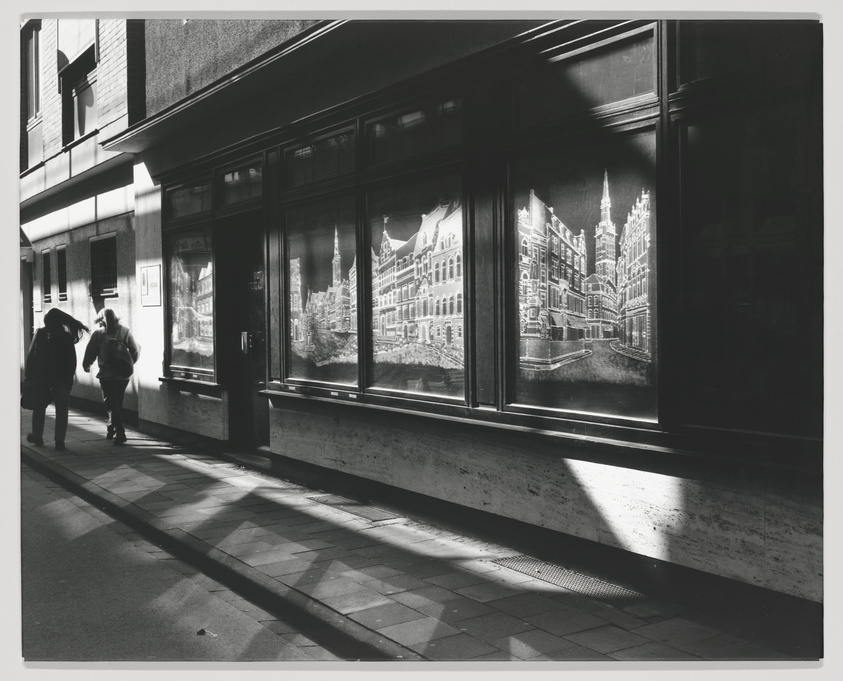 Black and white photograph showing two people walking past a building with large windows displaying artwork of historic buildings. The sunlight casts strong shadows on the sidewalk and the building's facade.