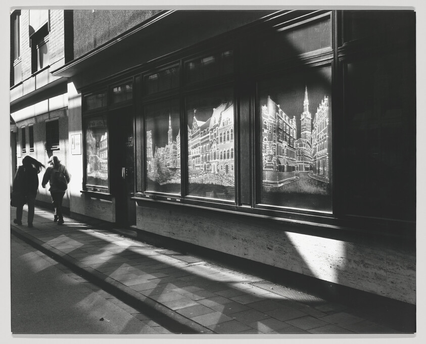 Two people walk past a storefront window showing illuminated architectural drawings and long sidewalk shadows.