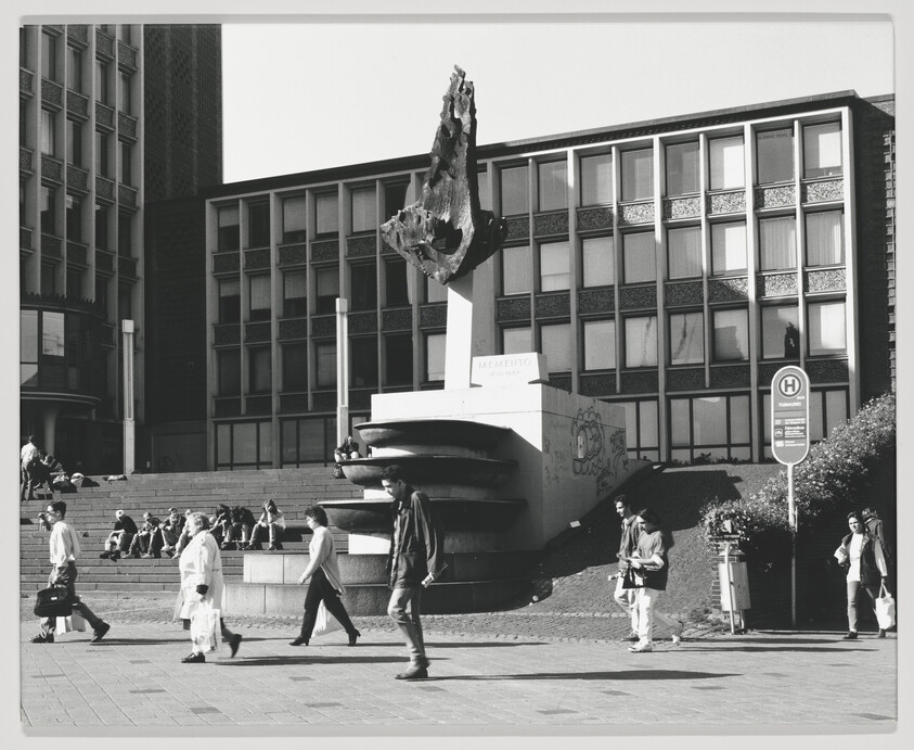 Large abstract sculpture on a pedestal in a city plaza with people walking and sitting.