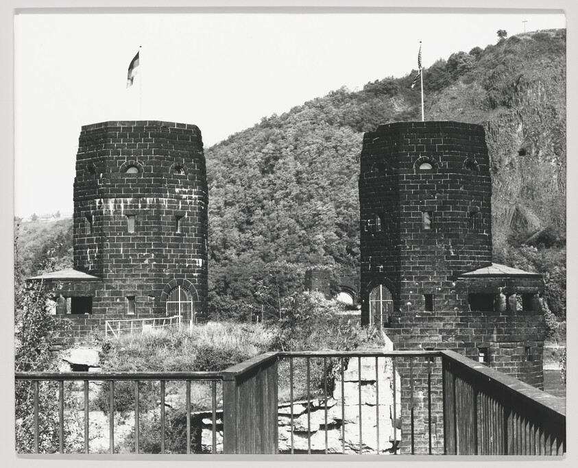 Two round stone bridge towers with flags stand by a river, seen from a railing.