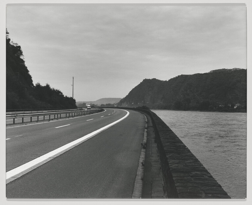A curved highway runs beside a river with guardrails and hills in the background.