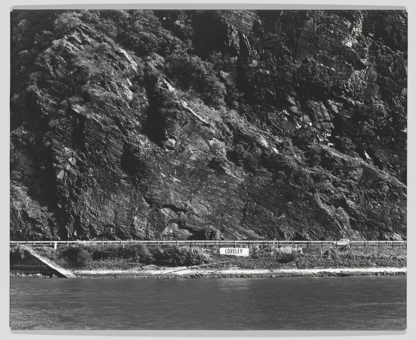 Steep rocky cliff looming above a riverside road with a sign reading Loreley.