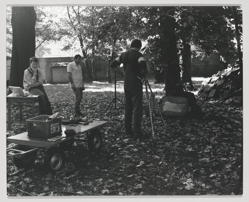 Three people set up camera equipment and lights under trees while one adjusts a tripod.