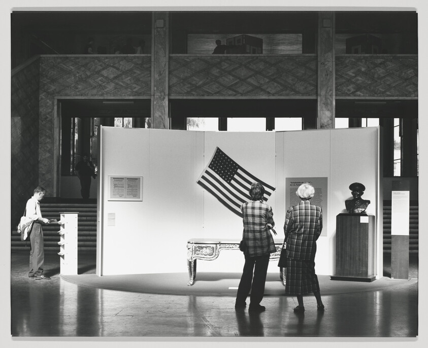 A black and white photo of an exhibition space with visitors. On the left, a person is reading an information panel. In the center, two individuals are observing an American flag on display. To the right, a bust is placed on a pedestal. The background features a large room with a herringbone-patterned floor and a balcony with people overlooking the area.