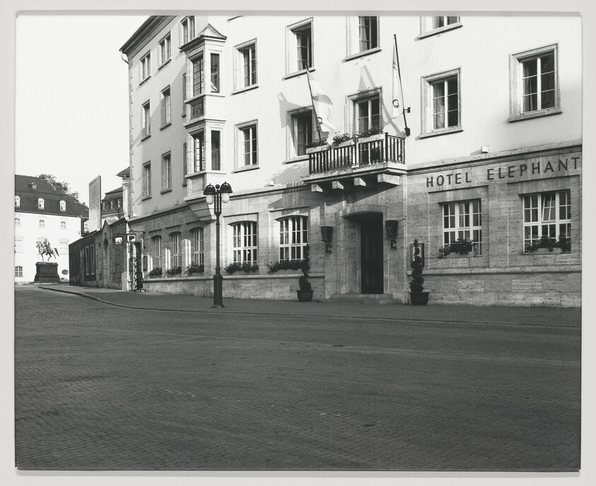Black and white photograph of a historic building with a sign reading "HOTEL ELEPHANT" on the facade. The hotel features classic architecture with multiple windows, flower boxes, and a balcony. A street lamp and a statue on a pedestal are visible on the cobblestone street in front of the hotel.
