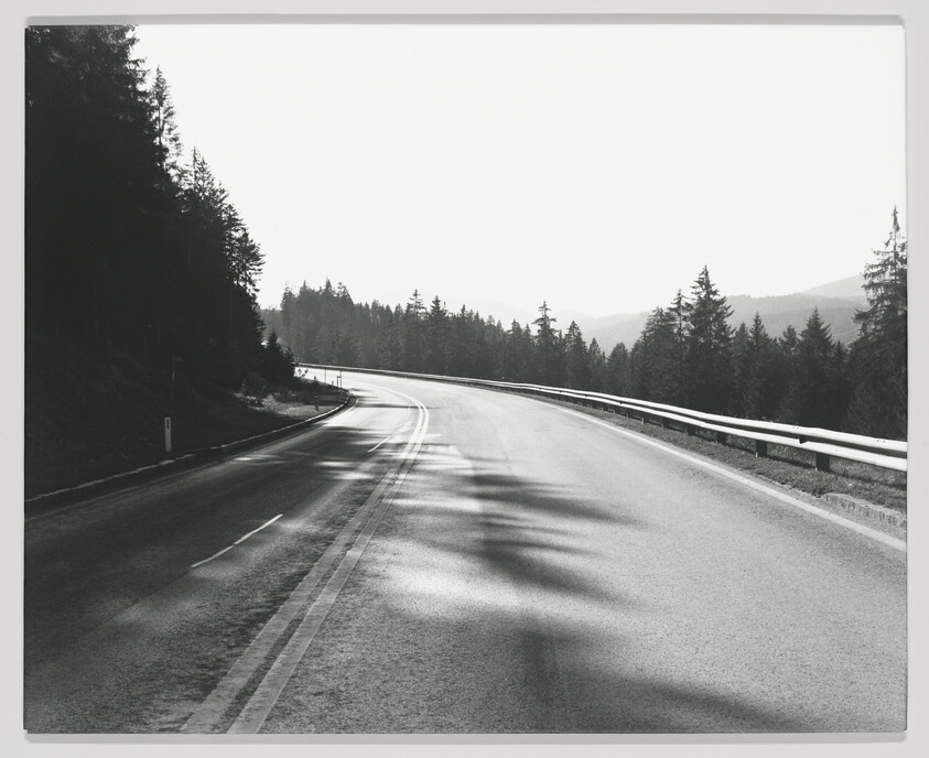 A black and white photograph of a curving road with a guardrail on the right, surrounded by dense forests and with mountains in the background. Shadows from an unseen source stretch across the road, suggesting a sunny day.