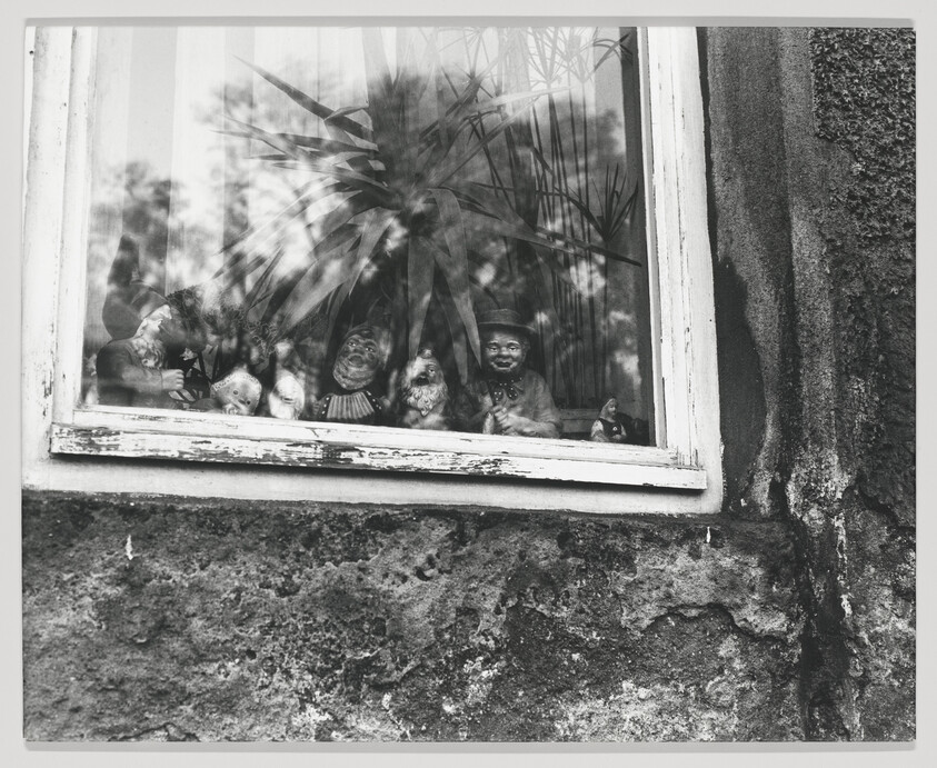 A black and white photograph showing a collection of vintage dolls and figurines displayed on a windowsill behind a glass pane. The reflection of trees and foliage can be seen overlaying the image of the dolls. The window frame is weathered, and the exterior wall beside the window has a rough, textured surface.