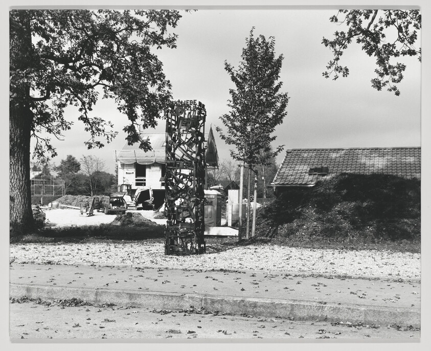 A black and white photograph depicting a rural scene with a large, intricate metal sculpture in the foreground. Behind the sculpture, construction equipment and a partially constructed building are visible, with a traditional house with a tiled roof in the background. Trees with autumn leaves frame the scene.
