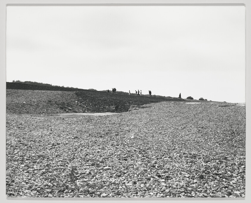 A black and white photograph depicting a pebble-covered beach leading up to a hill with several people standing at the top, under a clear sky.