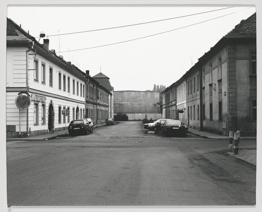 A black and white photograph of a quiet street scene with older buildings and parked cars, with a sign that reads "MOTOTECH" on the left side of the image.