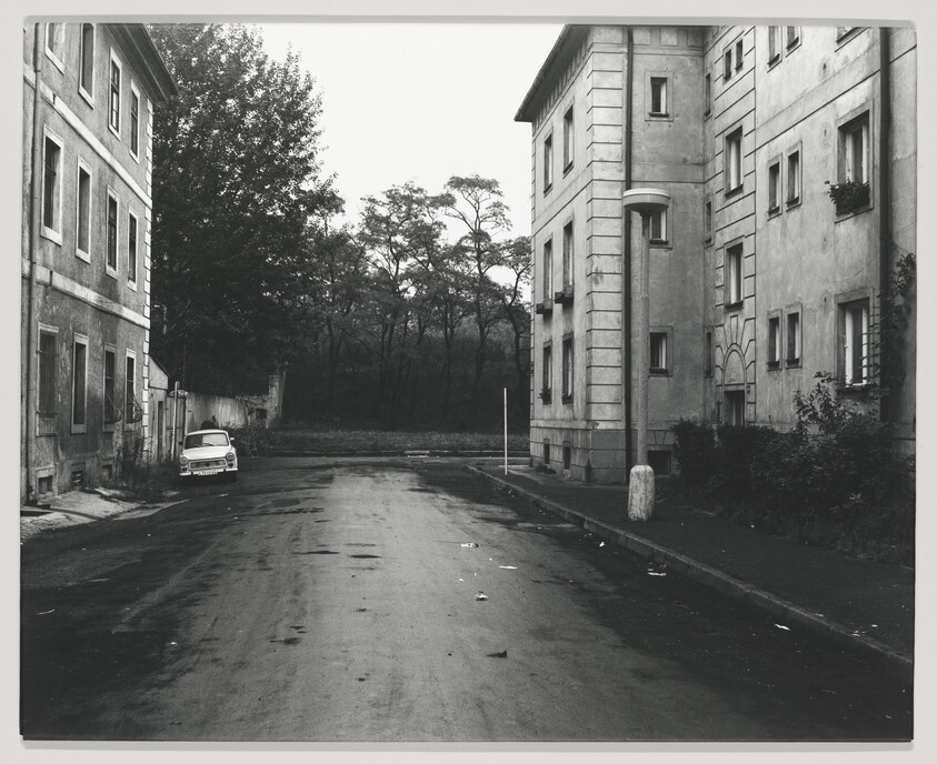 A black and white photograph depicting a deserted street flanked by old, weathered buildings with a single car parked on the side. Trees are visible in the background, suggesting the edge of a wooded area or park adjacent to the urban environment. The scene conveys a sense of quiet and abandonment.